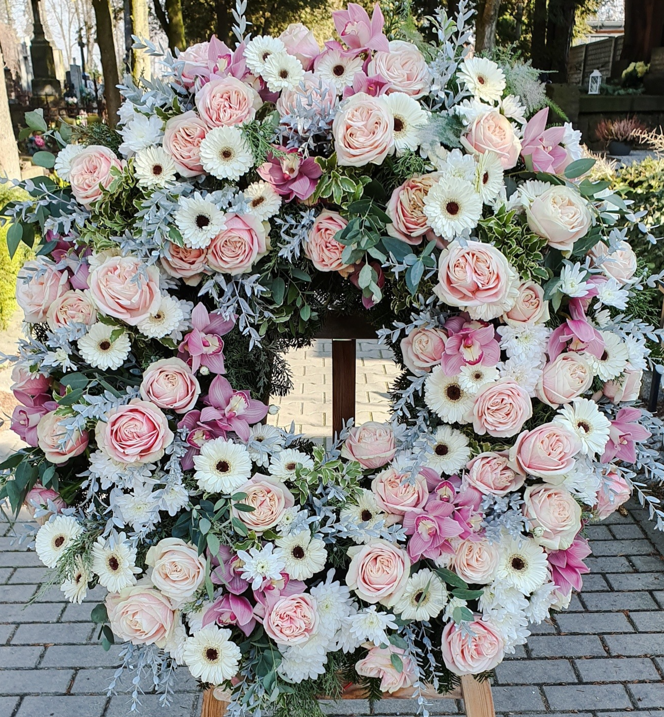 Bogaty, pastelowy wieniec funeralny ułożony z róż, storczyków, gerber i chryzantem. Duży okrągły wieniec z jasnoróżowych róż, białych gerber, storczyków oraz srebrzystych liści starca.