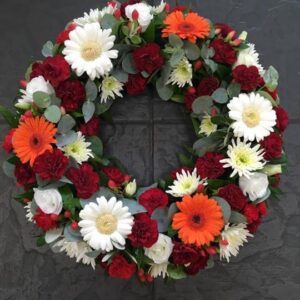 Okrągły wieniec rzymski z białych gerber, pomarańczowych chryzantem, bordowych goździków i eukaliptusa.