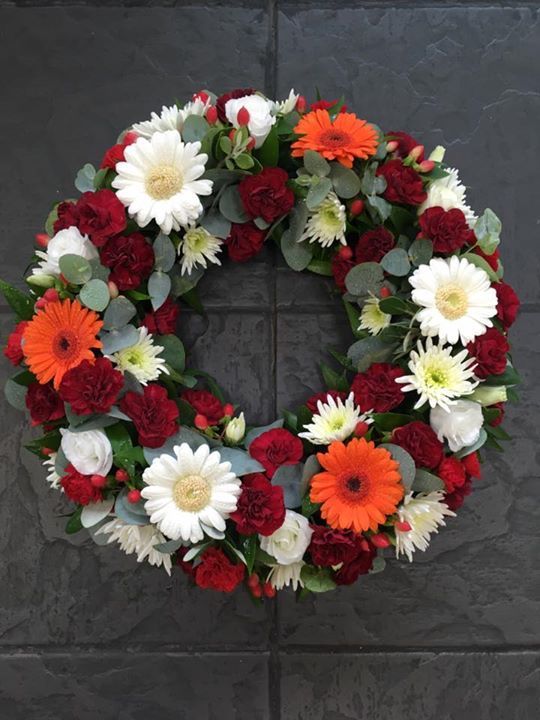 Okrągły wieniec rzymski z białych gerber, pomarańczowych chryzantem, bordowych goździków i eukaliptusa.