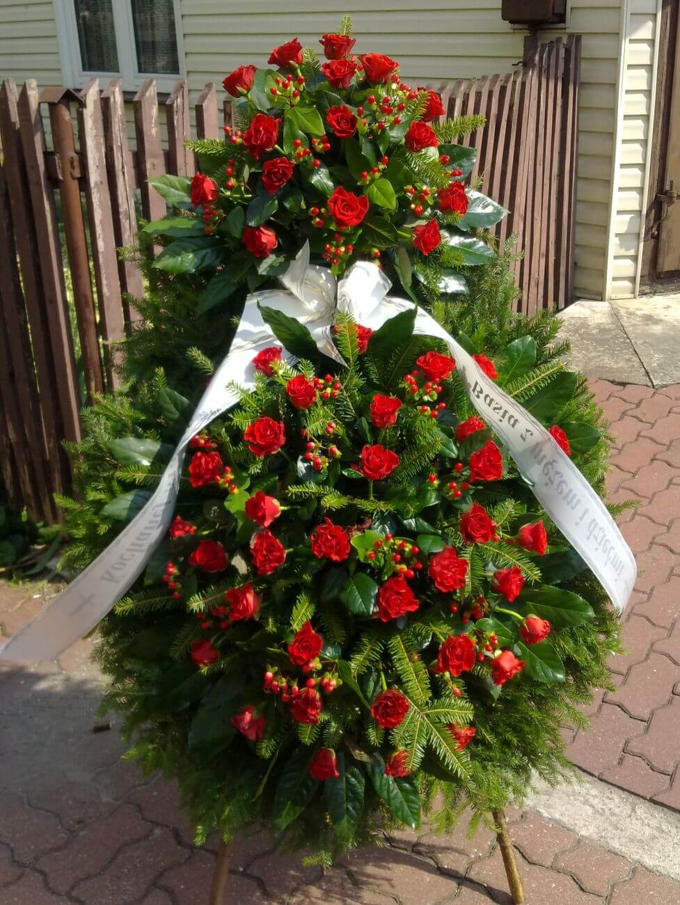 Ekskluzywny, dwupiętrowy wieniec funeralny z czerwonych róż – wyjątkowo dostojny symbol pamięci i hołdu. Dwupiętrowy wieniec pogrzebowy z czerwonych róż, dziurawca hypericum i białej szarfy na stojaku.